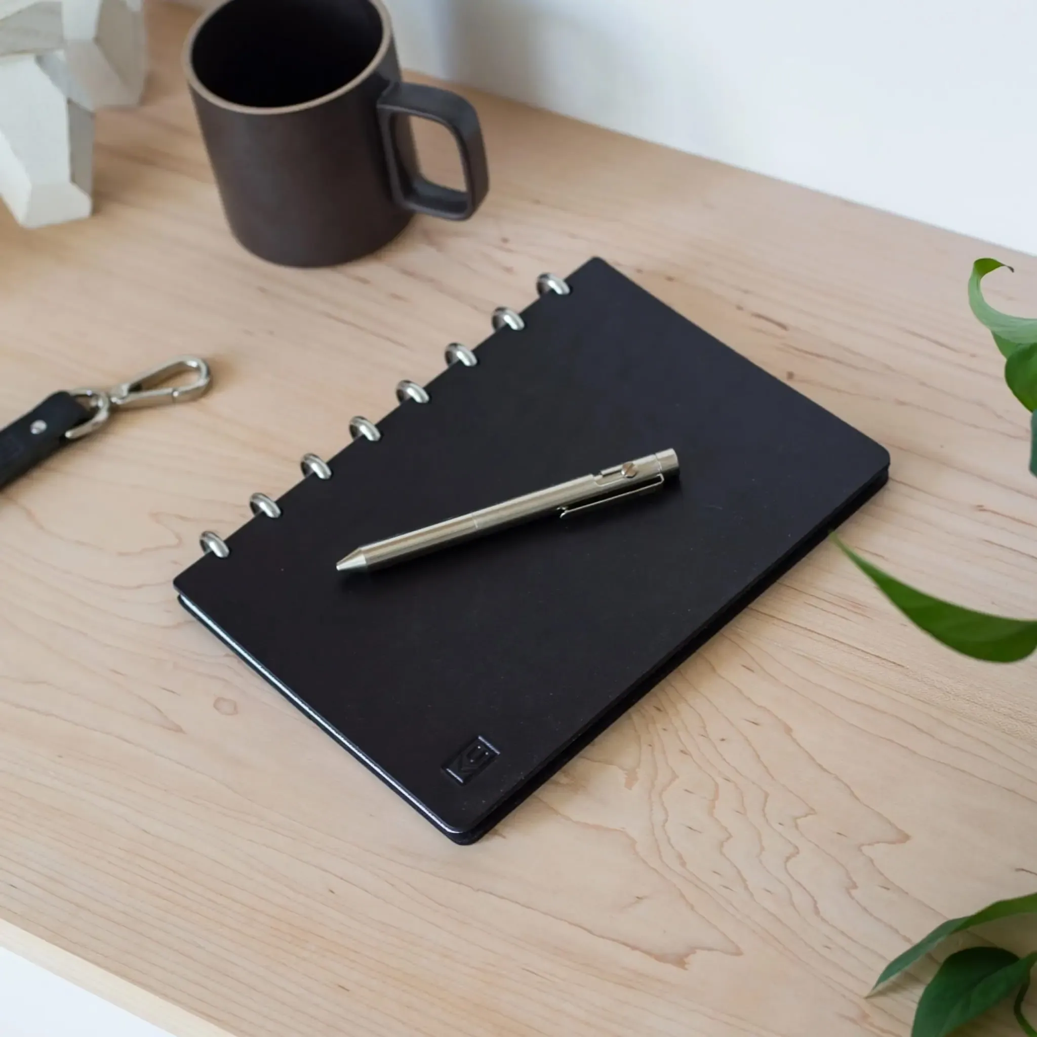 Black leather journal with silver steel disc binding resting on wooden shelf with machined steel pen.